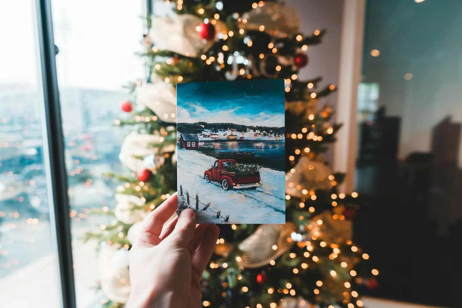 Ruka drží vytlačenú fotografiu pred ozdobeným vianočným stromčekom so svetielkami