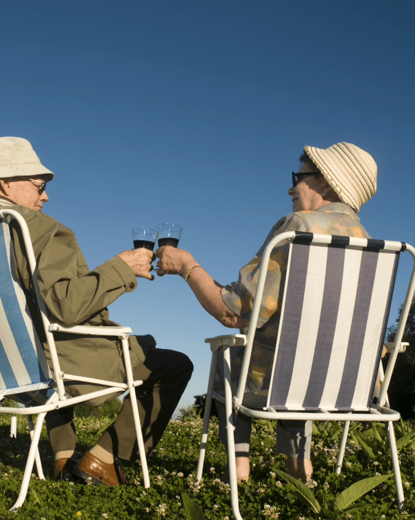 Starejši par se drži za roke in nazdravlja z vinom na stolih pod modrim nebom